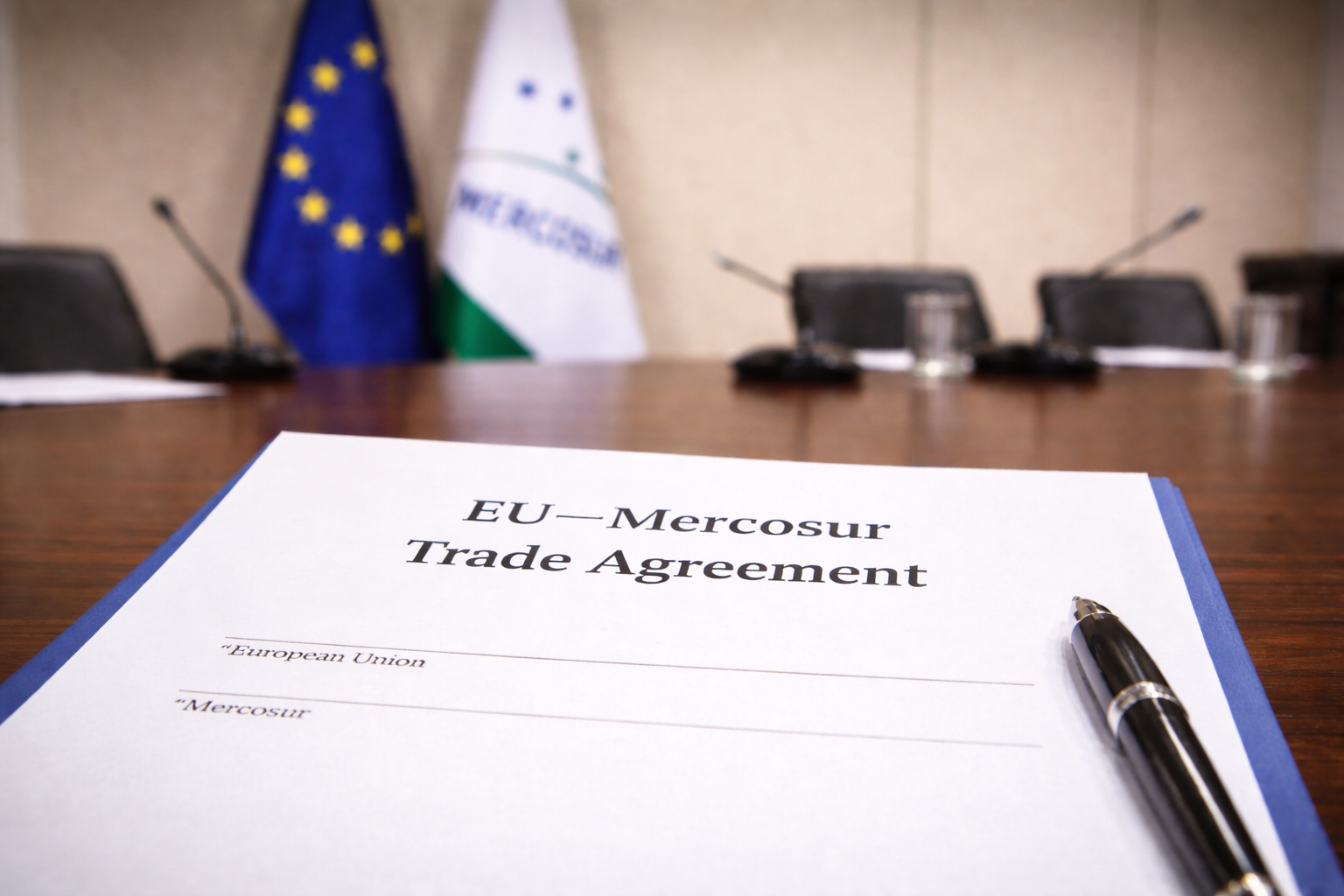 Documento del acuerdo comercial UE-Mercosur sin firmar sobre una mesa de negociación, con las banderas de la Unión Europea y Mercosur al fondo